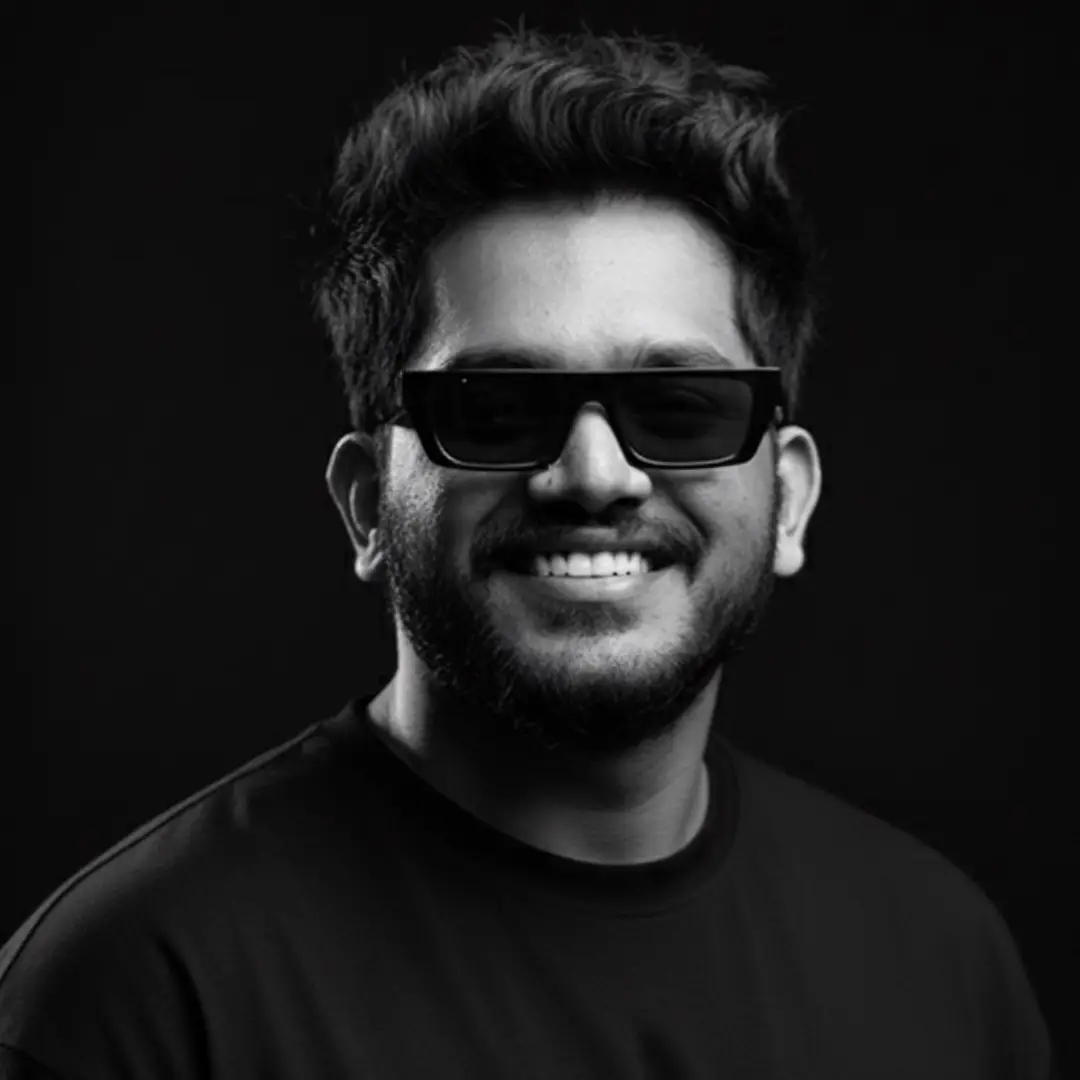 A black-and-white portrait of a smiling man with short, wavy hair, wearing dark sunglasses and a plain t-shirt, against a dark background